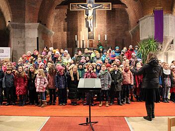 Kinderchor probt in einer Kirche für ein Weihnachtskonzert. Dirigentin steht vorne, viele Kinder bunt gekleidet. Hintergrund zeigt Kruzifix und Kerzen.