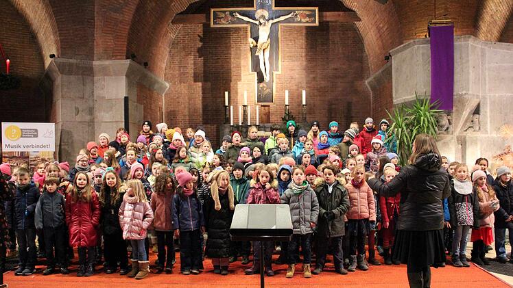 Kinderchor probt in einer Kirche für ein Weihnachtskonzert. Dirigentin steht vorne, viele Kinder bunt gekleidet. Hintergrund zeigt Kruzifix und Kerzen.