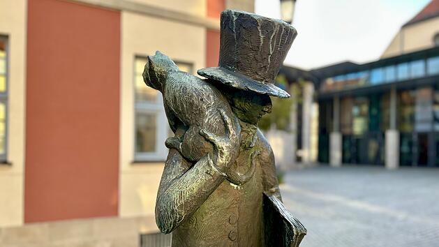 Eine Bronzestatue zeigt eine Figur mit Zylinder und Katze auf der Schulter, die ein Buch h&auml;lt. Sie steht vor einem Geb&auml;ude mit roten und beigen W&auml;nden und zieht sanftes Tageslicht an.