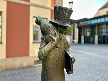 Eine Bronzestatue zeigt eine Figur mit Zylinder und Katze auf der Schulter, die ein Buch h&auml;lt. Sie steht vor einem Geb&auml;ude mit roten und beigen W&auml;nden und zieht sanftes Tageslicht an.