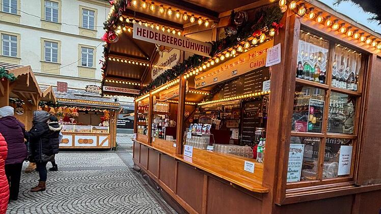 Eine Glühweinbude auf einem Weihnachtsmarkt bietet "Heidelbeer-Glühwein" an. Im Hintergrund sind weitere beleuchtete Stände zu sehen. Menschen in winterlicher Kleidung stehen davor.
