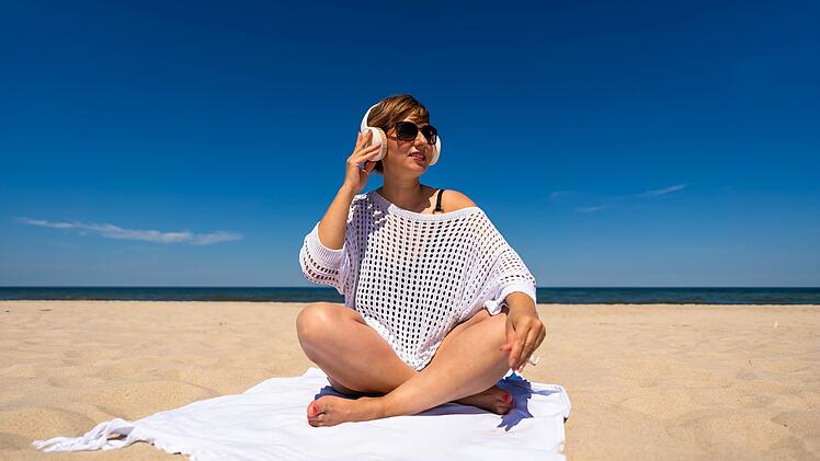 Sommerurlaub am Strand mit Musik. Schöne junge Frau in schwarzem Badeanzug und weißer Tunika hört Musik über Kopfhörer und sitzt im Schneidersitz auf einem Handtuch am weißen Sandstrand. Frontansicht
