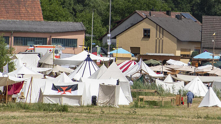 Ein mittelalterliches Lager mit zahlreichen weißen Zelten und Fahnen, umgeben von Bäumen und Gebäuden. Menschen in Gewändern bewegen sich zwischen den Zelten auf einer Wiese.