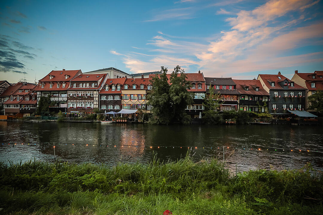 Bunte Fachwerkhäuser spiegeln sich im Fluss wider, umgeben von grünen Bäumen. Der Himmel ist blau mit rosa Wolken, die Szene wirkt idyllisch und friedlich.