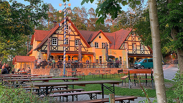 Ein malerischer fr&auml;nkischer Bierkeller bei Sonnenuntergang, umgeben von B&auml;umen. Es gibt Holzb&auml;nke und Tische im Freien, mit einem traditionellen Geb&auml;ude im Hintergrund.