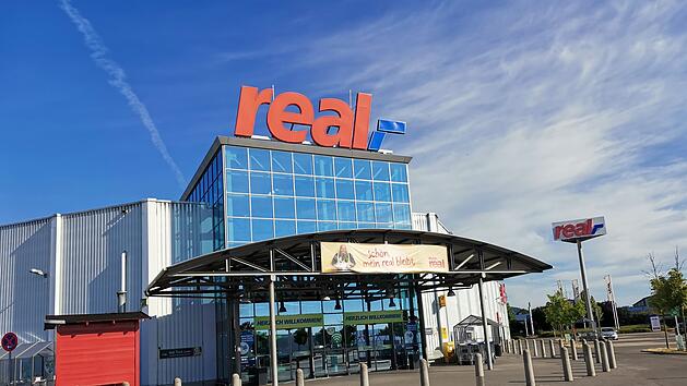 Ein "real"-Supermarkt mit blauem Glas-Eingang, rotem Logo und buntem Banner davor. Parkplatz und Himmel im Hintergrund. Baldige Er&ouml;ffnung einer Kaufland-Filiale in der Gegend ist geplant.
