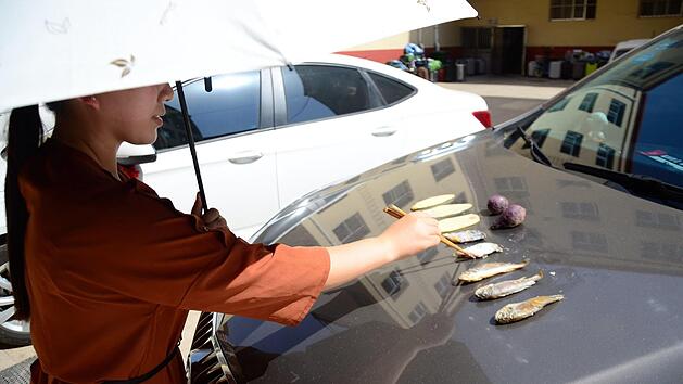 Eine Frau brät bei Temperaturen um die 40 Grad Celsius Fisch und Süßkartoffeln auf der aufgeheizten Motorhaube eines Autos. Foto: Wei Peng/SIPA Asia via ZUMA Wire/dpa