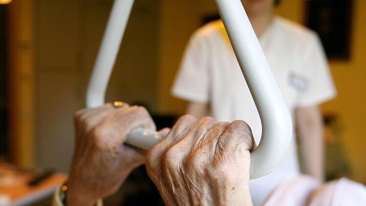 Eine Bewohnerin in einem Seniorenzentrum hält sich in ihrem Bett an einem Haltegriff fest, im Hintergrund steht eine Pflegekraft. Viele Menschen möchten lieber zuhause gepflegt werden. Aber wie lange bleibt das noch finanzierbar? Foto: dpa, Archiv, Angelika Warmuth