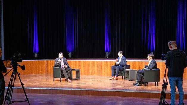 Moderator Frank Ebert (links) spricht in die Kamera, die Kandidaten Dominik Sauerteig (Mitte, rote Socken) und Christian Meyer (rechts, schwarze Socken) sitzen ihm im menschenleeren Kongresshaus gegen&uuml;ber. Die Aufzeichnung des Talks ist online bei itv-coburg.de zu finden. Foto: Simone Bastian