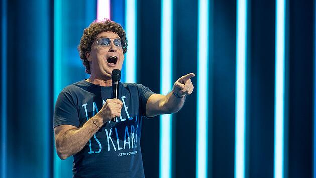 Ein Mann steht mit Mikrofon auf einer B&uuml;hne, tr&auml;gt ein T-Shirt mit blauer Aufschrift. Im Hintergrund leuchten vertikale blaue Lichtstreifen.