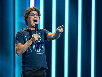 Ein Mann steht mit Mikrofon auf einer B&uuml;hne, tr&auml;gt ein T-Shirt mit blauer Aufschrift. Im Hintergrund leuchten vertikale blaue Lichtstreifen.