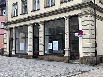 Ein historisches Geb&auml;ude mit gro&szlig;en Fenstern, in denen Schilder h&auml;ngen. Ein verkehrsberuhigtes Stra&szlig;enschild steht vorne. Das Umfeld ist ruhig und gepflastert.