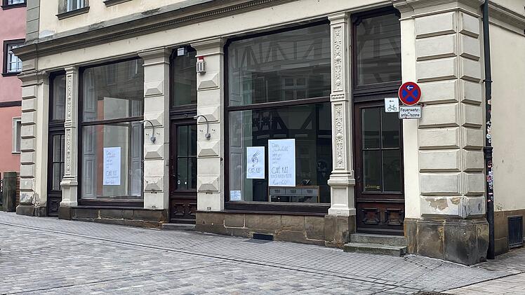 Ein historisches Geb&auml;ude mit gro&szlig;en Fenstern, in denen Schilder h&auml;ngen. Ein verkehrsberuhigtes Stra&szlig;enschild steht vorne. Das Umfeld ist ruhig und gepflastert.