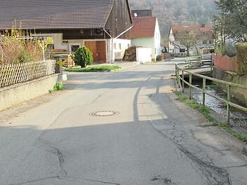 Der Blick aus Richtung Hundshaupten in die Ortsmitte von Oberzaunsbach zeigt den schlechten Fahrbahnzustand und die erneuerungsbedürftige Bachmauer.
