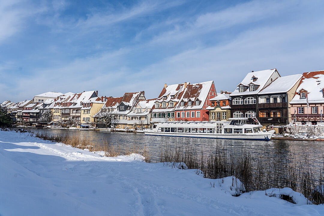 Verschneite H&auml;userreihe am Wasser in Bamberg. Ein wei&szlig;es Boot f&auml;hrt auf dem Fluss, w&auml;hrend dichte Schneewehen den Rand bedecken. Klarer blauer Himmel erg&auml;nzt das Winterwunder.