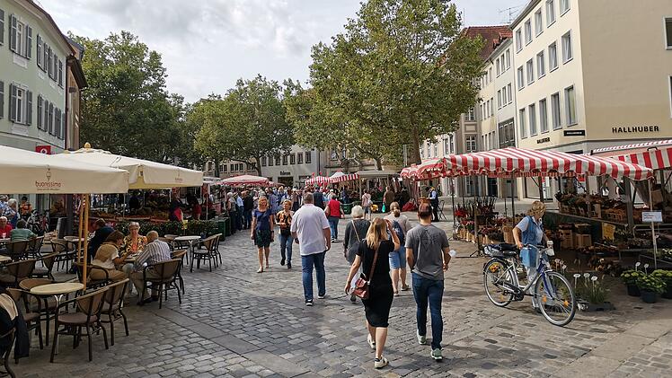 Ein belebter Marktplatz in Bamberg mit St&auml;nden und Caf&eacute;s, umgeben von alten Geb&auml;uden. Menschen schieben Fahrr&auml;der und bummeln durch die Stra&szlig;e.