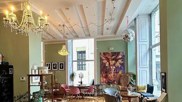 Gem&uuml;tliches Caf&eacute; mit Holzboden, pastellgr&uuml;nen W&auml;nden, eleganten Kronleuchtern, Kunst an den W&auml;nden und verschiedenen Sitzbereichen. Einladende Atmosph&auml;re mit gro&szlig;en Fenstern.