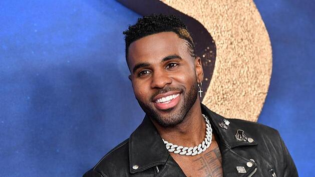 Ein Mann in schwarzer Lederjacke l&auml;chelt vor einem blauen Hintergrund mit goldenen Details.