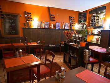 Ein gemütliches, orangefarbenes italienisches Restaurant mit Holztischen, Weinflaschen an der Wand und Tafeln mit dem Menü. Blumen und Pflanzen dekorieren den Raum.