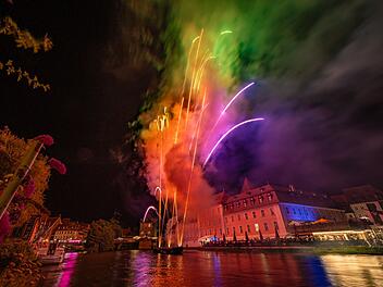 Farbenfrohes Feuerwerk über einem Fluss bei Nacht, reflektiert auf dem Wasser, Häuser im Hintergrund beleuchtet. Stimmungsvoll und festlich, passend zur Bamberger Sandkerwa.