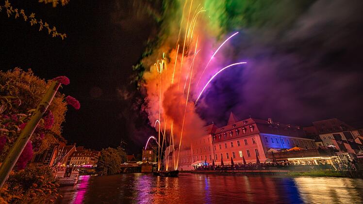Farbenfrohes Feuerwerk über einem Fluss bei Nacht, reflektiert auf dem Wasser, Häuser im Hintergrund beleuchtet. Stimmungsvoll und festlich, passend zur Bamberger Sandkerwa.