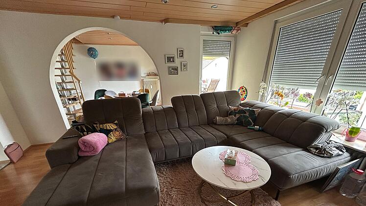 Ein modernes Wohnzimmer mit einer großen, grauen Eckcouch und einem runden Couchtisch mit Spitzendecke. Im Hintergrund ist ein Essbereich und eine Treppe zu sehen.