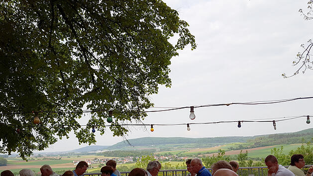Menschen sitzen an Holztischen unter einem Baum auf einem fr&auml;nkischen Bierkeller. Im Hintergrund gibt es eine sch&ouml;ne Aussicht auf Felder und H&uuml;gel. Lichter sind &uuml;ber den Tischen aufgeh&auml;ngt.