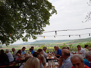Menschen sitzen an Holztischen unter einem Baum auf einem fr&auml;nkischen Bierkeller. Im Hintergrund gibt es eine sch&ouml;ne Aussicht auf Felder und H&uuml;gel. Lichter sind &uuml;ber den Tischen aufgeh&auml;ngt.