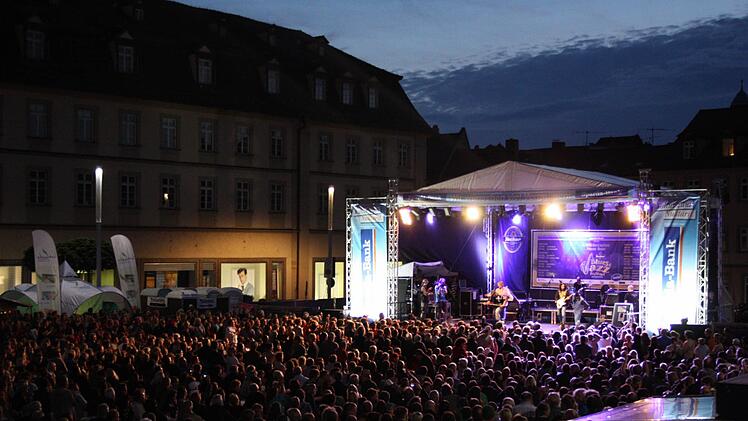 Ein lebhaftes Blues- & Jazzfestival bei Nacht in Bamberg. Eine große Menschenmenge genießt Live-Musik auf einer beleuchteten Bühne. Banner und Lichter schaffen eine feierliche Atmosphäre.