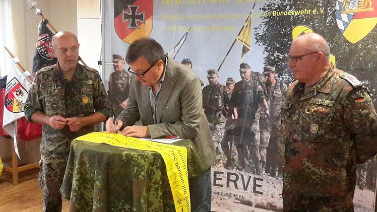 Riedenbergs Bürgermeister Roland Römmelt unterschreibt auf dem gelben Band. Die zwei Meter lange Banderole wird später symbolisch einer Einheit übergeben, die in den Auslandseinsatz geht. Interessierte Beobachter sind die Reservisten Johann Schmidt (links) und Karl Veit (rechts).Foto: Rolf Pralle