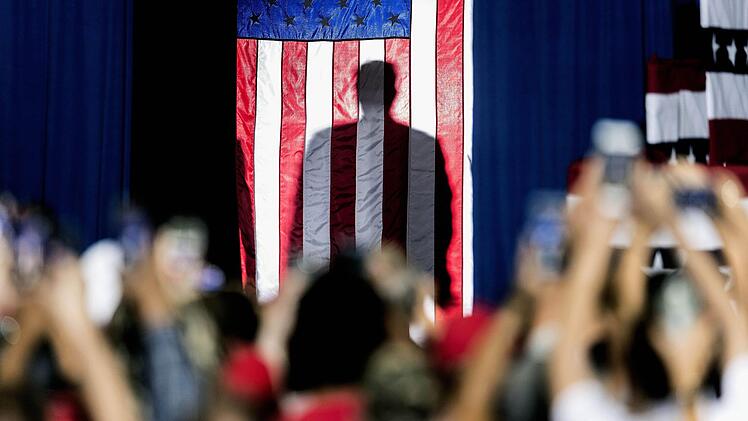 Donald J. Trumps Schatten hinter der amerikanischen Flagge, als er sich auf seinen Auftritt bei einer "Keep America Great"-Kundgebung im Santa Ana Star Center vorbereitet (Rio Rancho, USA, 2019).