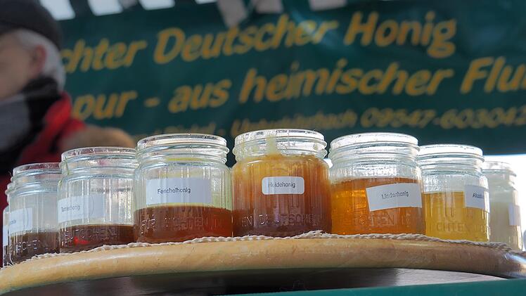 Verschiedene Honiggl&auml;ser auf einem Tisch. Im Hintergrund ein Schild mit der Aufschrift &bdquo;Echter Deutscher Honig&ldquo;. Eine Person ist teilweise sichtbar.