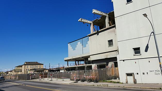 Ein teilweise abgerissenes Geb&auml;ude neben einer Stra&szlig;e. Bauarbeiten laufen, Bauz&auml;une sind aufgestellt. Im Hintergrund ein weiteres Geb&auml;ude. Der Himmel ist klar und blau.