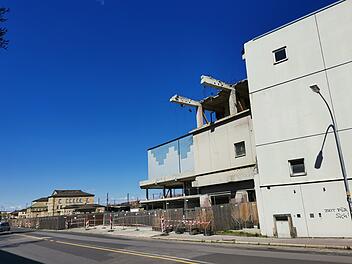 Ein teilweise abgerissenes Gebäude neben einer Straße. Bauarbeiten laufen, Bauzäune sind aufgestellt. Im Hintergrund ein weiteres Gebäude. Der Himmel ist klar und blau.