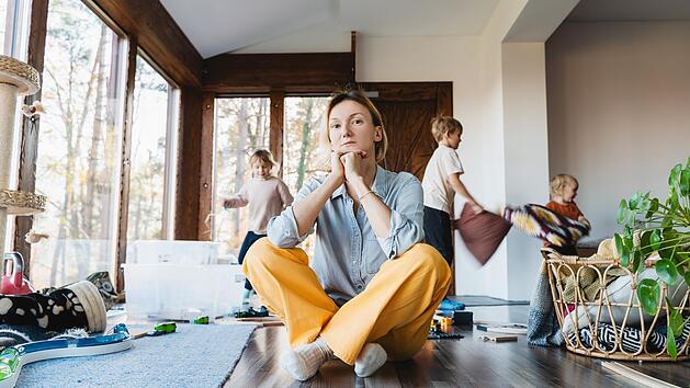Frustrierende Frau allein kämpft Burnout mit Kindern. Gestresste Mutter sitzt auf dem Boden in der Mitte des Spielzeugs, während die Kinder frech um sie herum im Zimmer laufen. Familie zu Hause mit Chaos, Unordnung. Alleinerziehend, Stress, Kinder.Frustrating woman alone struggling burnout with kids. Stressed out mother sitting on floor in middle of toys while children naughty running around her at room. Family home with chaos, mess.