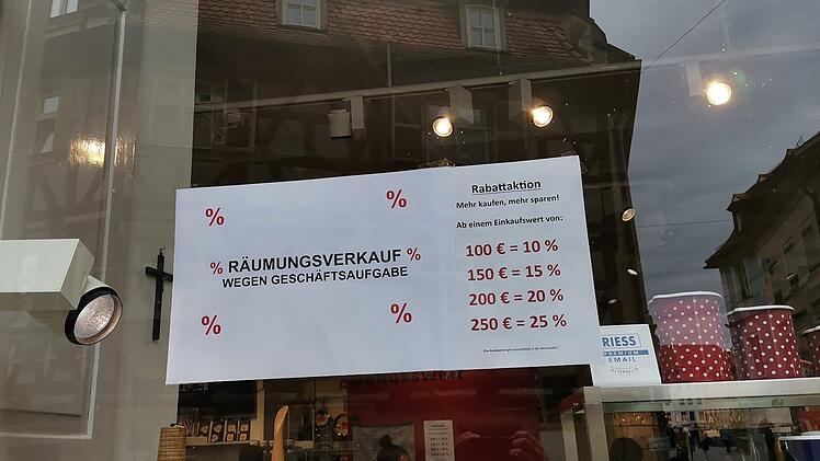 Ein Schaufenster mit einem Schild für Räumungsverkauf wegen Geschäftsschließung. Rabattaktionen bieten 10-25% Ermäßigung. Reflektionen zeigen die Altstadt von Bamberg.