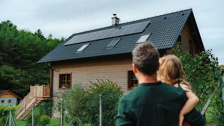 Rear view of dad holding her little girl in arms and looking at their house with installed solar panels. Alternative energy, saving resources and sustainable lifestyle concept.
