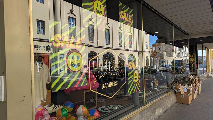 Ein Schaufenster zeigt "Sale"-Schilder mit einem Smiley und "love". Im Inneren sind bunte Artikel und Kartons zu sehen. Der Shop in Bamberg bleibt geöffnet, während andere schließen.