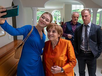 Charu Borscht (Farina Flebbe, links), die junge Frau des zweiten Bestatters im Ort, macht ein Selfie mit Angela Merkel (Katharina Thalbach). Ehemann und Bestatter Borscht (Bernhard Sch&uuml;tz) sowie Merkels Personensch&uuml;tzer Mike (Tim Kalkhof, rechts) schauen zu.