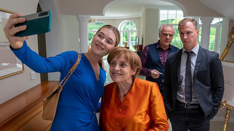 Charu Borscht (Farina Flebbe, links), die junge Frau des zweiten Bestatters im Ort, macht ein Selfie mit Angela Merkel (Katharina Thalbach). Ehemann und Bestatter Borscht (Bernhard Sch&uuml;tz) sowie Merkels Personensch&uuml;tzer Mike (Tim Kalkhof, rechts) schauen zu.