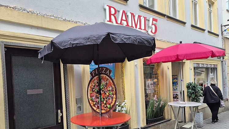 Vor einem Restaurant steht ein rot-grauer Tisch mit einem Pizzabild und Schirmen. Ein Gesch&auml;ft namens "Rami 5" ist im Hintergrund sichtbar. Passant geht vorbei.