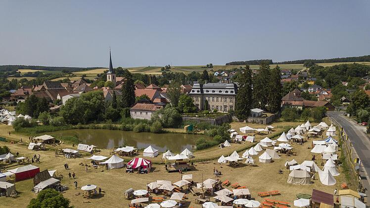Ein mittelalterliches Lager mit zahlreichen Zelten und Besuchern ist in Burgwindheim aufgebaut, nahe eines Teiches und eines historischen Gebäudes, umgeben von idyllischer Landschaft.