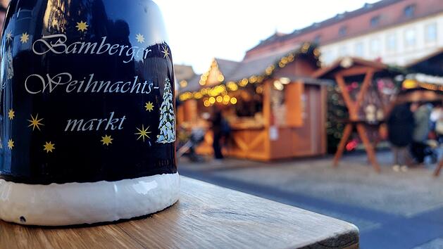 Ein Bamberger Weihnachtsmarktbecher im Vordergrund, dahinter festlich geschm&uuml;ckte St&auml;nde. Typische vorweihnachtliche Atmosph&auml;re mit Lichtern und Holzh&uuml;tten.