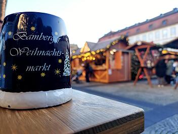 Ein Bamberger Weihnachtsmarktbecher im Vordergrund, dahinter festlich geschmückte Stände. Typische vorweihnachtliche Atmosphäre mit Lichtern und Holzhütten.