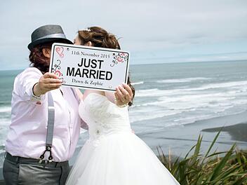 Mit einem Schild "Just Married" stehen Dawn und Zephie aus Singapur im Jahr 2015 nach ihrer Hochzeit am Strand von Auckland (Neuseeland). Neuseeland hat 2013 die gleichgeschlechtliche Ehe legalisiert und ist seitdem ein Magnet für Ausländer, die heiraten wollen. Foto: Amanda Wignell Photography /dpa