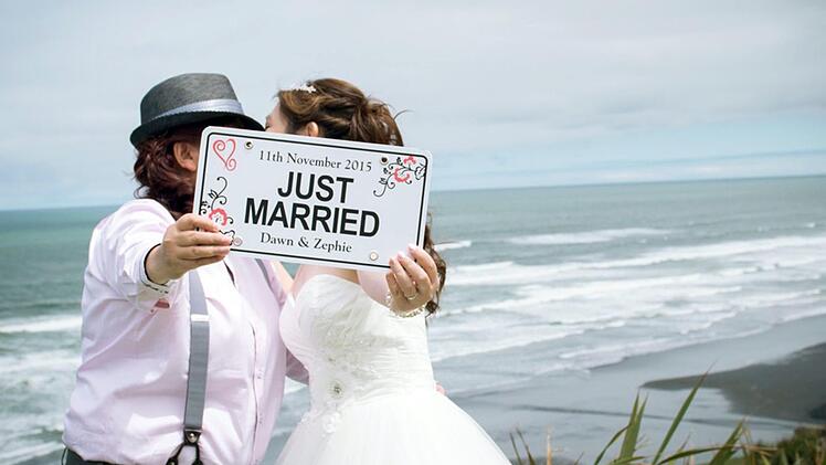Mit einem Schild "Just Married" stehen Dawn und Zephie aus Singapur im Jahr 2015 nach ihrer Hochzeit am Strand von Auckland (Neuseeland). Neuseeland hat 2013 die gleichgeschlechtliche Ehe legalisiert und ist seitdem ein Magnet für Ausländer, die heiraten wollen. Foto: Amanda Wignell Photography /dpa