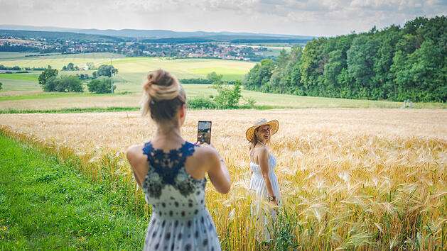 Zwei Frauen im Sommerkleid in einem goldenen Weizenfeld, eine fotografiert die andere. Der Hintergrund zeigt H&uuml;gel und W&auml;lder unter einem leicht bew&ouml;lkten Himmel.