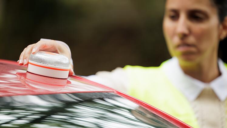 Ab 2026 dürfen Autofahrer und Autofahrerinnen in Spanien bei Unfällen oder Pannen auf Autobahnen kein Warndreieck mehr aufstellen. Pflicht wird dann ein spezielles Blinklicht. Young woman with reflective vest, places the V16 emergency light beacon on the roof of the damaged vehicle. Help flash, mandatory to replace triangles in Europe. DGT Spain.Junge Frau mit Warnweste platziert die Notfallleuchte V16 auf dem Dach des beschädigten Fahrzeugs. Hilfe-Blinklicht, in Europa als Ersatz für Warndreiecke vorgeschrieben. DGT Spanien.