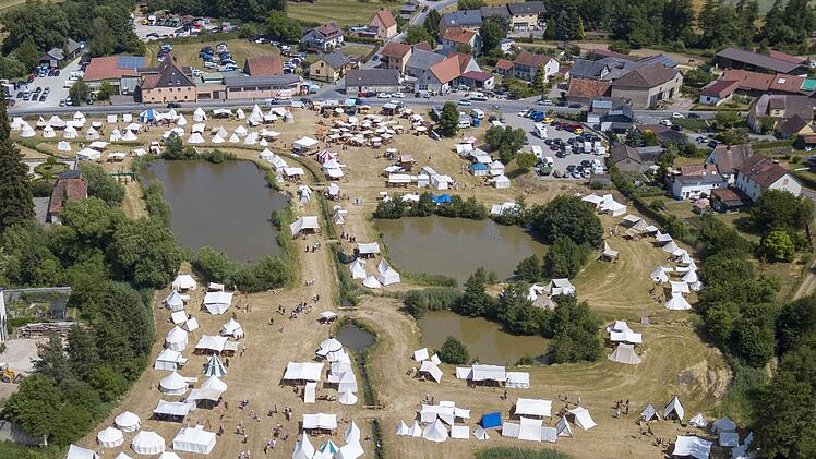 Zelte umgeben von Teichen in ländlicher Gegend. Menschen in historischen Gewändern, Lager verteilt, dient als Touristenattraktion. Häuser im Hintergrund, viele Besucher. Veranstaltung Burgwindheim.