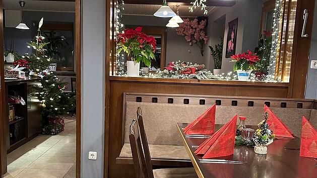 Ein gem&uuml;tlich dekorierter Raum mit Weihnachtsthema. Ein gedeckter Tisch mit roten Servietten, Weihnachtsschmuck und einem Weihnachtsbaum mit Geschenken im Hintergrund.